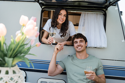 Cheerful couple with wine glasses looking at camera near camper van