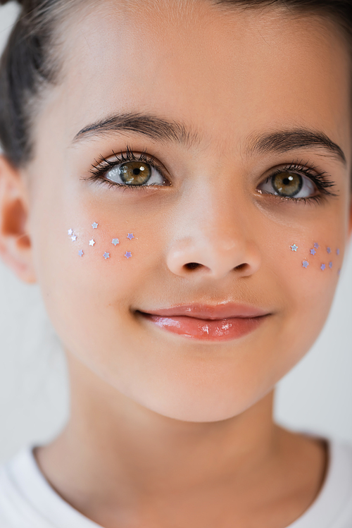 close up view of cheerful girl with hazel eyes and sparkling glitter stars on cheeks isolated on grey