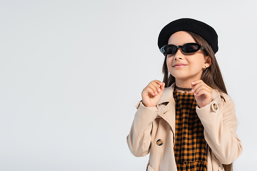 happy girl in stylish trench coat and sunglasses smiling isolated on grey