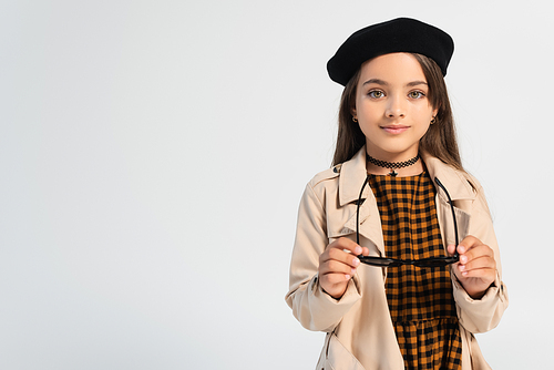 happy girl in stylish trench coat and beret holding sunglasses isolated on grey