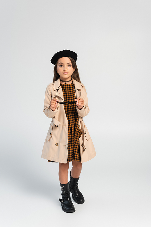 full length of cute girl in stylish trench coat and beret holding sunglasses on grey