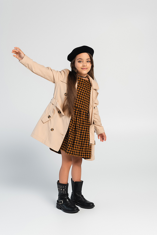 full length of cheerful girl in stylish trench coat and beret gesturing while standing on grey