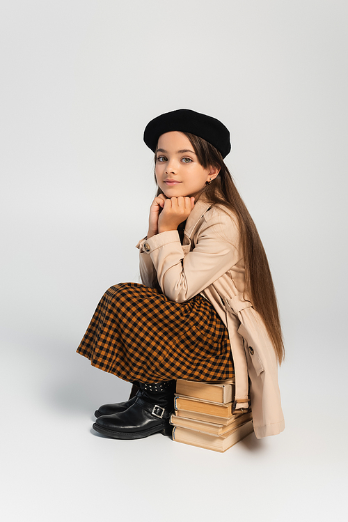 full length of cute kid in stylish autumnal outfit and beret sitting on stacked books on grey