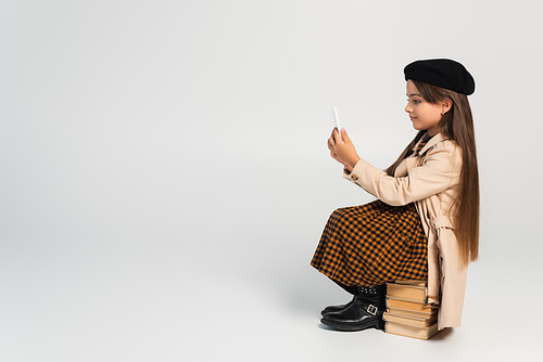 full length of cute kid in stylish autumnal outfit and beret sitting on stacked books and using smartphone on grey