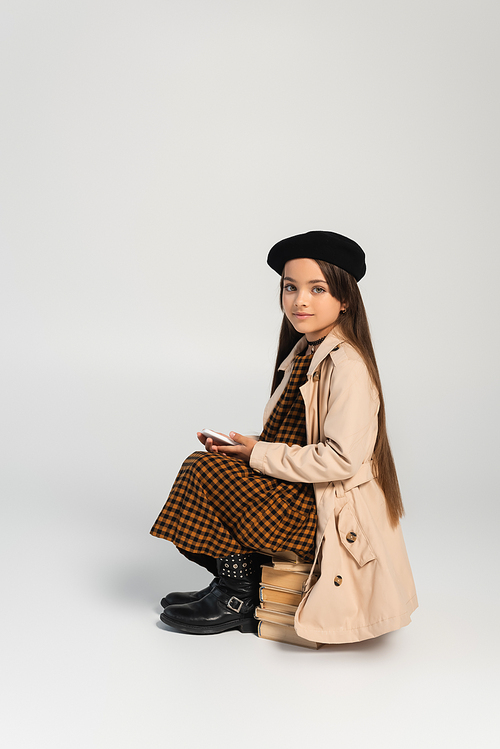 full length of cute kid in stylish autumnal outfit and beret sitting on stacked books and holding smartphone on grey