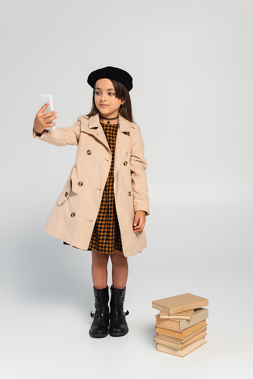 full length of cute kid in stylish autumnal outfit and beret taking selfie near stacked books on grey