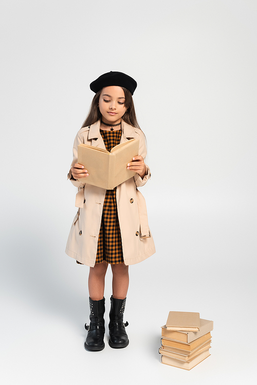 full length of cute kid in stylish autumnal outfit and beret reading book on grey