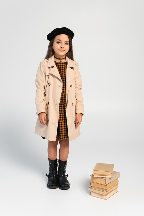 full length of smiling kid in stylish autumnal outfit and beret standing near stacked books on grey