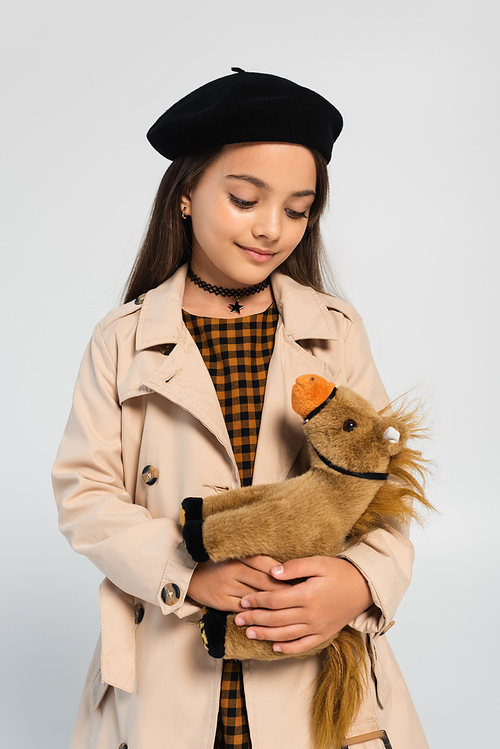 cheerful girl in stylish trench coat and beret standing with toy horse isolated on grey