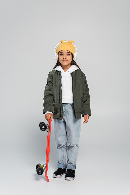 full length of cheerful girl in headphones and trendy autumnal outfit holding penny board on grey