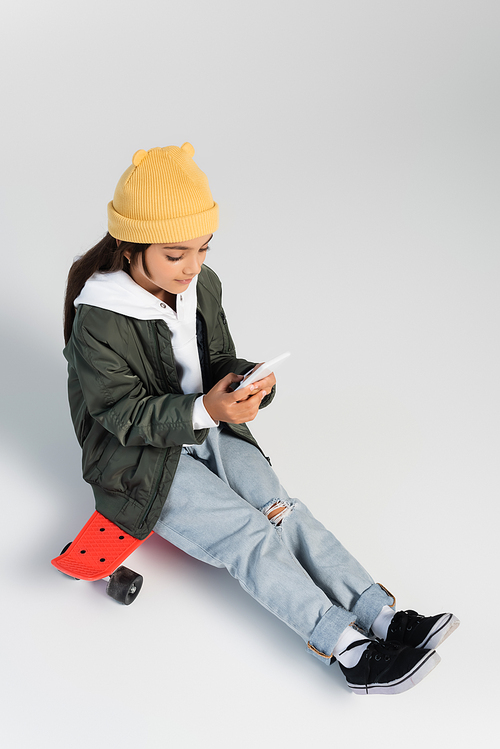 high angle view of cute girl in trendy autumnal outfit using smartphone and sitting on penny board on grey