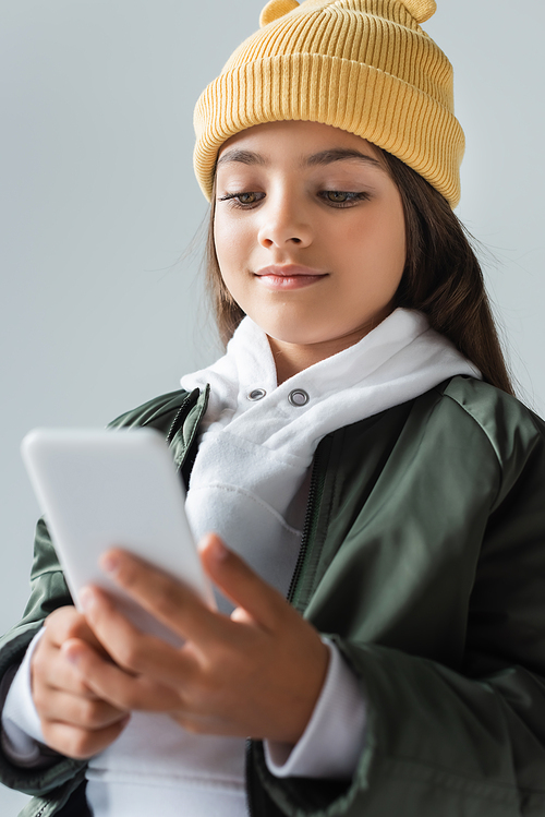 cheerful kid in trendy autumnal outfit and beanie hat using smartphone isolated on grey