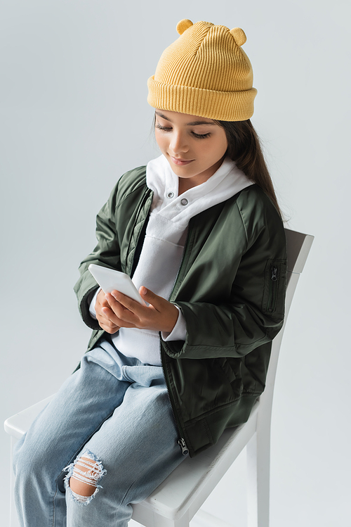 high angle view of cheerful kid in trendy autumnal outfit sitting on high chair and using smartphone isolated on grey