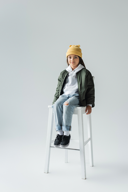 full length of cute girl in trendy autumnal outfit sitting on high chair on grey