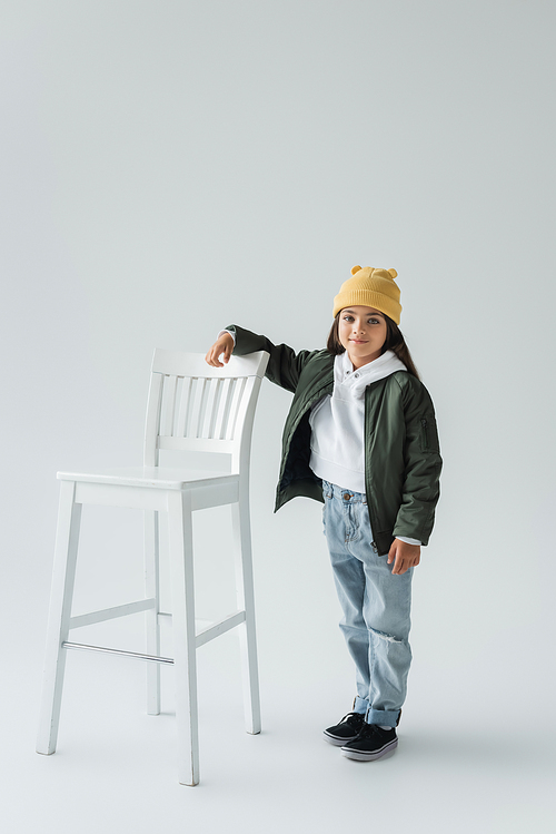 full length of cheerful girl in trendy autumnal outfit and beanie hat standing near high chair on grey