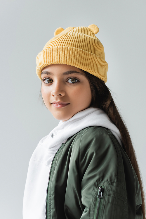 portrait of adorable girl in yellow beanie hat and bomber jacket looking at camera isolated on grey