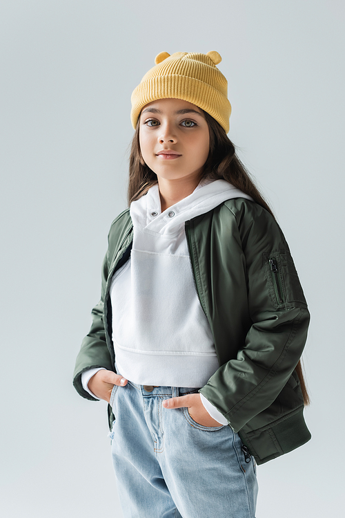 portrait of adorable girl in yellow beanie hat and stylish autumnal outfit posing with hands in pockets isolated on grey