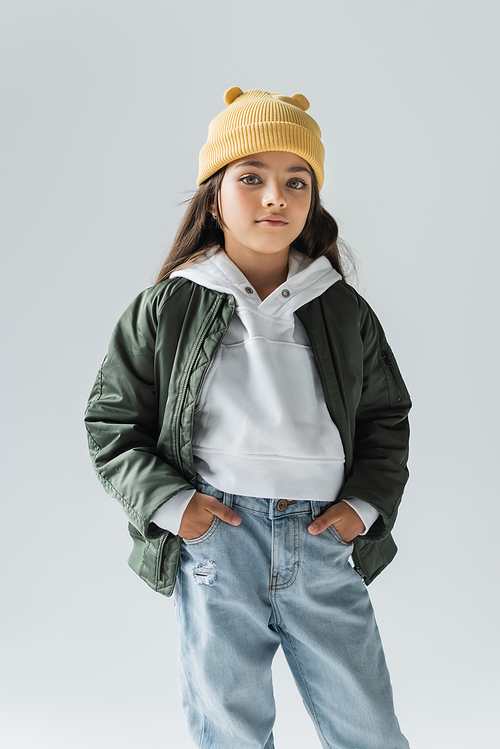 portrait of cute kid in yellow beanie hat and stylish autumnal outfit posing with hands in pockets isolated on grey