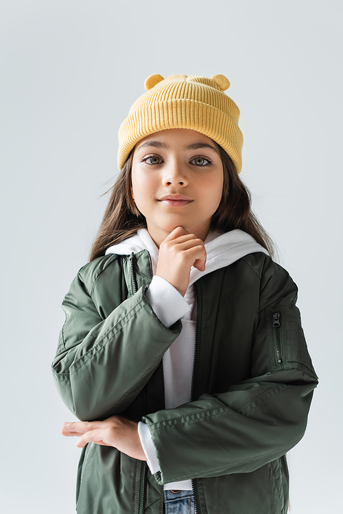 cheerful girl in yellow beanie hat and bomber jacket looking at camera isolated on grey