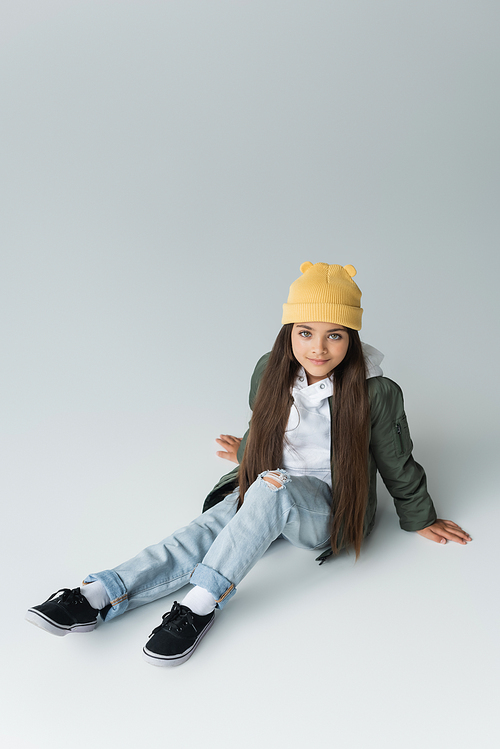 high angle view of adorable girl in trendy autumnal outfit and beanie hat sitting on grey