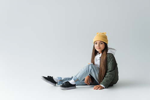 full length of adorable girl in trendy autumnal outfit and beanie hat sitting on grey