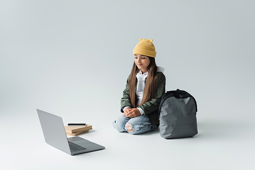 full length of adorable girl in trendy autumnal outfit looking at laptop while sitting near smartphone on books and backpack on grey