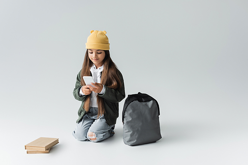 full length of adorable girl in trendy autumnal outfit using smartphone while sitting near books and backpack on grey