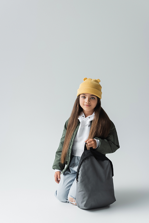 full length of cheerful kid in trendy autumnal outfit and beanie hat holding backpack on grey