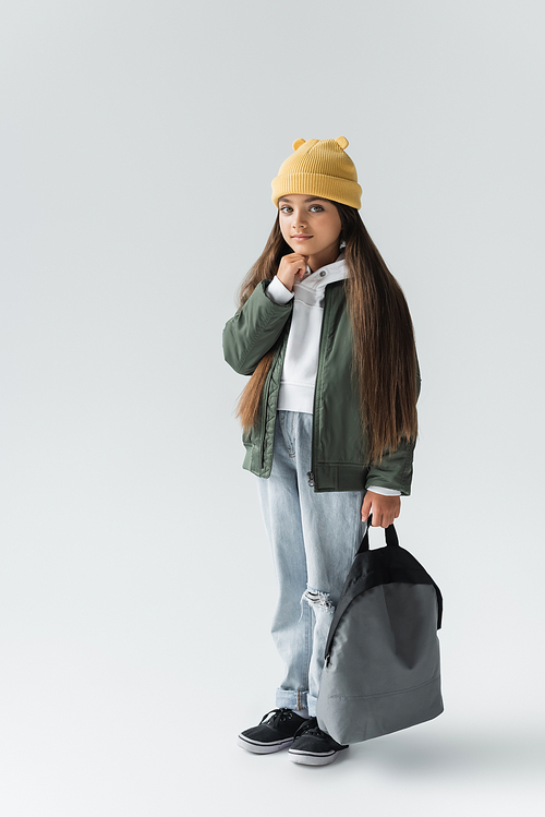 full length of adorable girl in trendy autumnal outfit and beanie hat holding backpack on grey