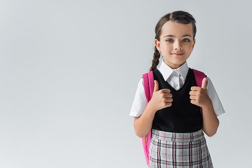 cheerful schoolgirl with backpack standing in school uniform and showing thumbs up isolated on grey