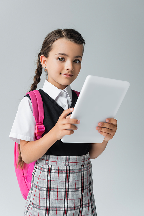 happy and cute schoolgirl in uniform holding digital tablet while smiling isolated on grey