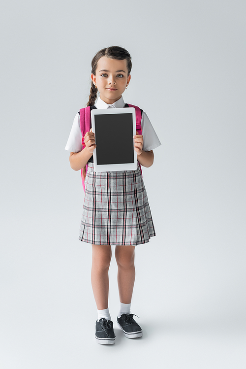 full length of cute schoolgirl in uniform holding digital tablet with blank screen on grey