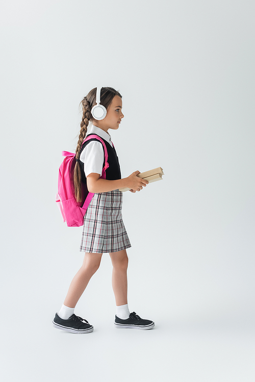 side view of girl in school uniform and wireless headphones walking with books on grey