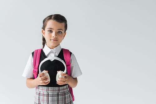 schoolgirl in uniform standing with backpack and holding wireless headphones isolated on grey