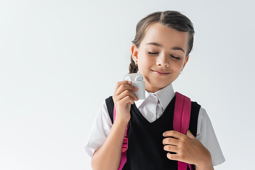 pleased schoolgirl with closed eyes holding earphone case isolated on grey