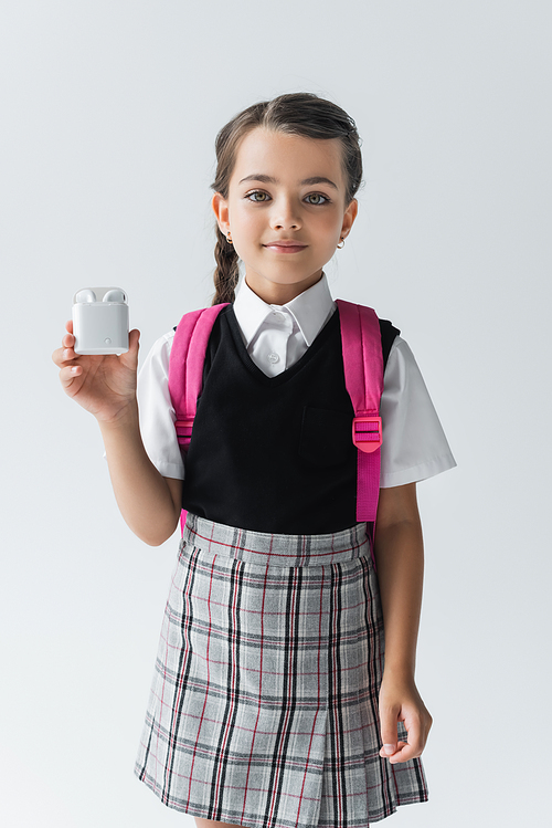 cheerful schoolgirl in uniform standing with backpack and holding earphone case isolated on grey