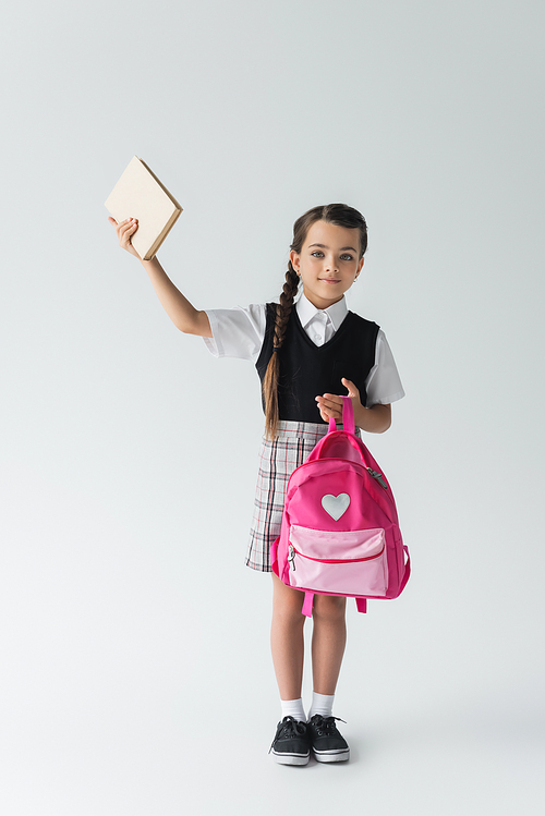 full length of cute schoolgirl in uniform holding pink backpack and book above head on grey