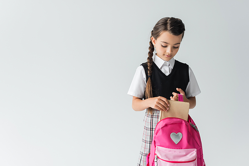 cute schoolgirl in uniform putting book in pink backpack isolated on grey