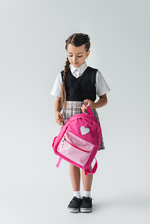 full length of cute schoolgirl in uniform holding pink backpack while standing on grey