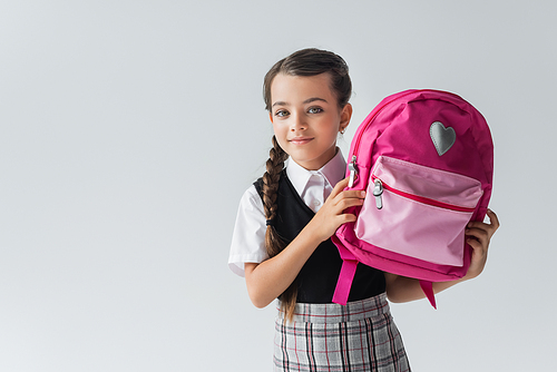 cute schoolgirl in uniform holding pink backpack and smiling isolated on grey