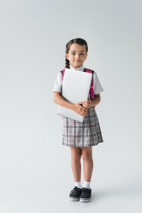 full length of schoolgirl in uniform standing with backpack and laptop on grey