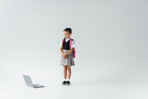 full length of schoolgirl in uniform standing with backpack and looking at laptop on grey