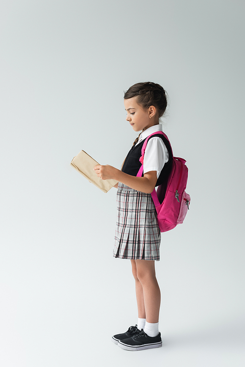 side view of smart schoolgirl in uniform reading book and standing and reading book on grey