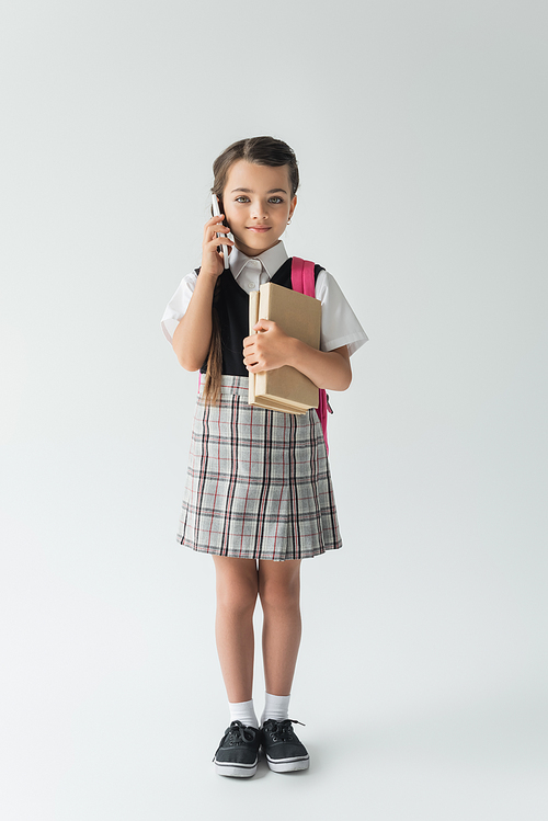 full length of smiling schoolgirl talking on smartphone and holding books on grey