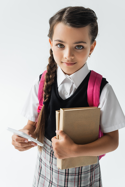 high angle view of smiling schoolgirl holding smartphone and books isolated on grey