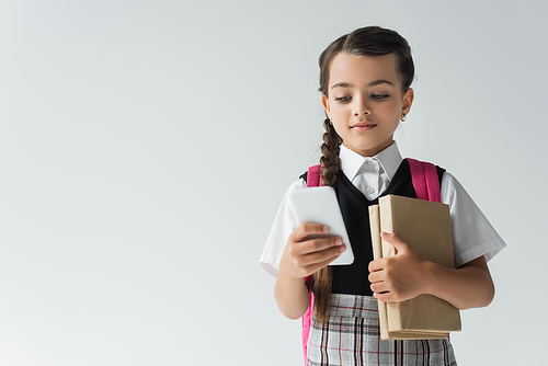 adorable schoolgirl in uniform using smartphone and holding books isolated on grey