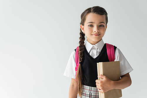 adorable schoolgirl in uniform holding books and smiling isolated on grey