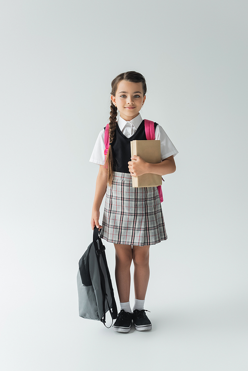 full length of adorable schoolgirl in uniform holding books and backpack on grey