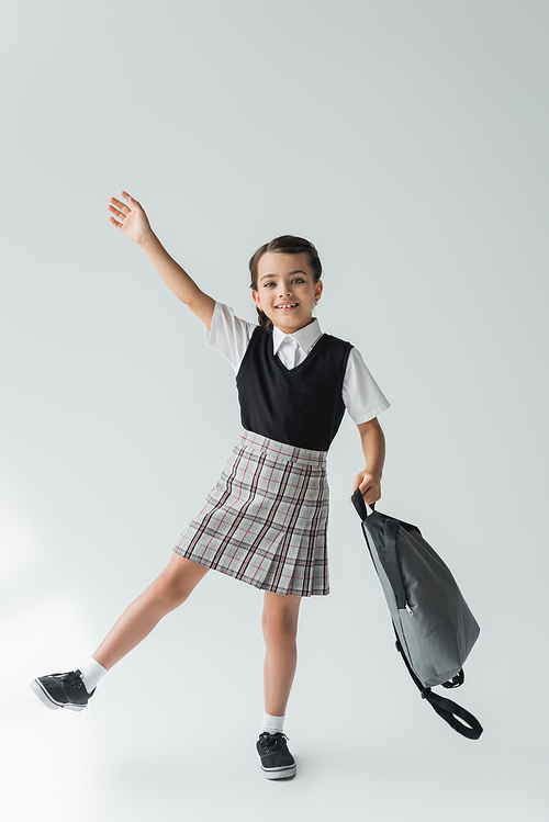 full length of adorable and cheerful schoolgirl in uniform holding backpack on grey