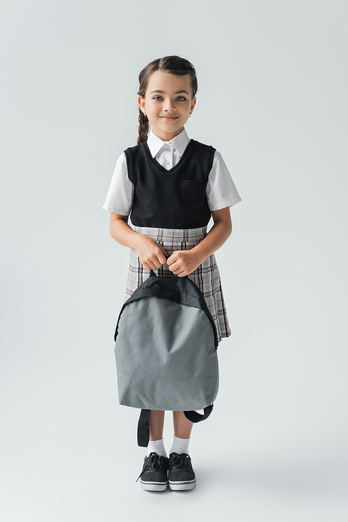 full length of adorable and happy schoolgirl in uniform holding backpack on grey